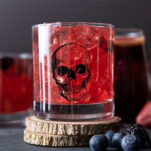 May include: A glass filled with red liquid and ice cubes, decorated with a black skull graphic. The glass is sitting on a wooden coaster.