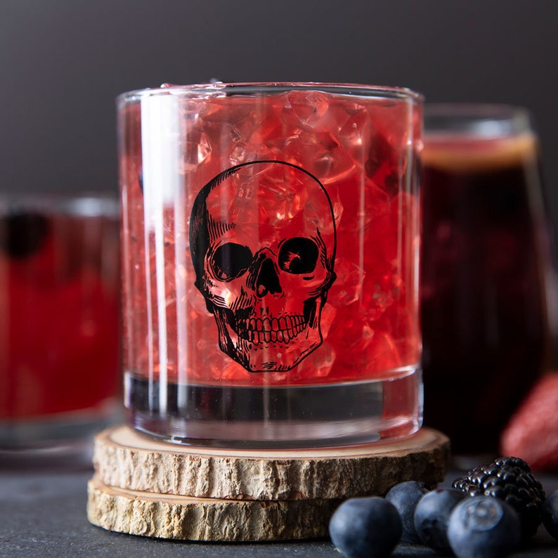 May include: A glass filled with red liquid and ice cubes, decorated with a black skull graphic. The glass is sitting on a wooden coaster.