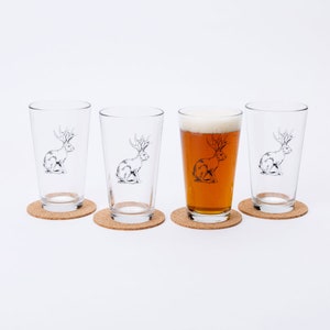 May include: Four clear glass pint glasses with a black and white illustration of a rabbit with antlers on each. One glass is filled with a light brown beer. Each glass is sitting on a round cork coaster.