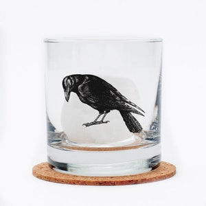 May include: A clear glass tumbler with a black and white illustration of a crow on the side. The tumbler is sitting on a cork coaster.