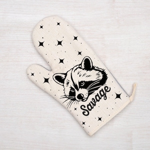 Puede incluir: Manopla de horno color crema con un gráfico de mapache en blanco y negro y la palabra "Savage". La manopla está decorada con formas de estrellas negras. Se adjunta un lazo para colgar. Tema de cocina y cocina.