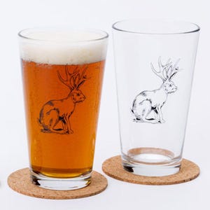 May include: Two clear glass pint glasses with a black and white illustration of a rabbit with antlers. The glass on the left is filled with amber beer. Both glasses are sitting on round cork coasters.
