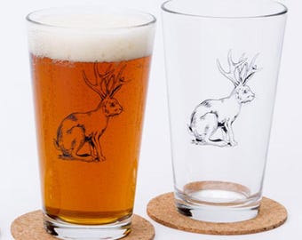 May include: Two clear glass pint glasses with a black and white illustration of a rabbit with antlers. The glass on the left is filled with amber beer. Both glasses are sitting on round cork coasters.