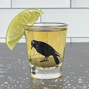 May include: A clear shot glass with a black crow design on the side. The glass is filled with a light yellow liquid and a lime wedge is resting on the rim.
