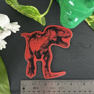 May include: Red vinyl sticker of a black and white illustration of a Tyrannosaurus Rex dinosaur.