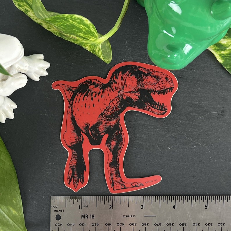 May include: Red vinyl sticker of a black and white illustration of a Tyrannosaurus Rex dinosaur.