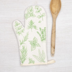 May include: Off-white oven mitt with green botanical illustrations of herbs, including basil, rosemary, and thyme. A wooden spoon rests beside the mitt. The mitt has a loop for hanging.
