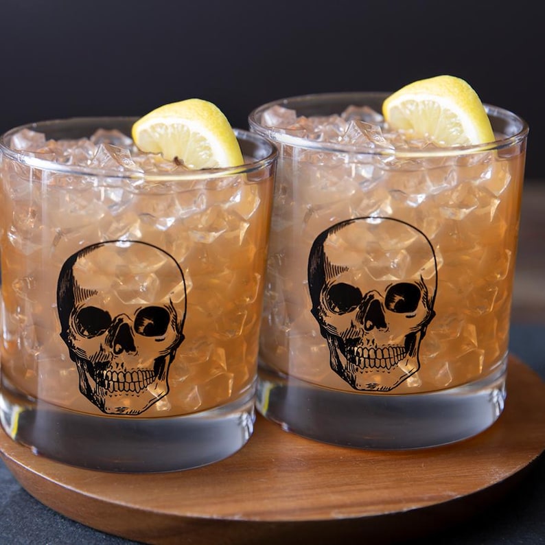 May include: Two clear glass tumblers with a skull design. The glasses are filled with ice and a brown liquid. A lemon wedge is in each glass.