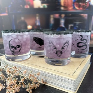 May include: Four clear glass tumblers with black rims filled with a purple liquid. Each glass features a different black and white illustration: a skull, a crow, a bat, and a snake.