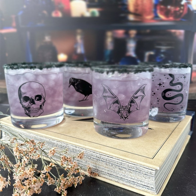 May include: Four clear glass tumblers with black rims filled with a purple liquid. Each glass features a different black and white illustration: a skull, a crow, a bat, and a snake.