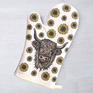 May include: An oven mitt with a Highland cow design, surrounded by yellow sunflowers on a white background. The cow has brown fur and large horns. The mitt has a loop for hanging and is made of fabric.