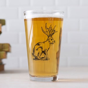 May include: A clear glass pint glass with a black and white illustration of a rabbit with antlers. The glass is filled with a light amber colored beer.