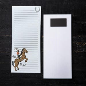 May include: White notepad with lined paper and a black magnet. The notepad features a cartoon of a person riding a brown horse, with the words "Yee Haw" in black. A horseshoe is at the top. The notepad is on a dark wooden surface.