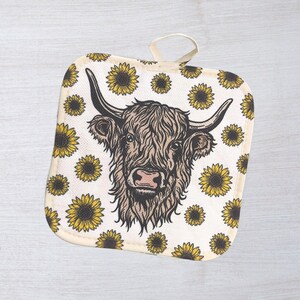 May include: Square beige pot holder with a loop for hanging. The pot holder features a detailed illustration of a Highland cow's head, surrounded by yellow sunflowers. The cow has long, shaggy brown fur and large horns.