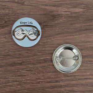 Op de afbeelding: Een lichtblauwe knoop met de tekst "Slope Life" en een afbeelding van een skibril met een berglandschap. De knoop is rond en heeft een speld aan de achterkant. De knoop heeft een diameter van ongeveer 3,2 cm.