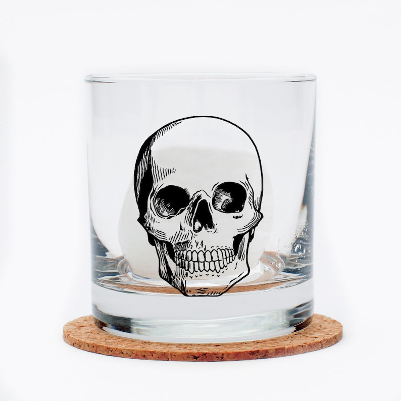 May include: A clear glass tumbler with a black and white skull graphic. The tumbler is sitting on a cork coaster.
