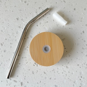 May include: A silver metal straw and a wooden lid with a small hole in the center. The lid is light brown and has a natural wood grain.