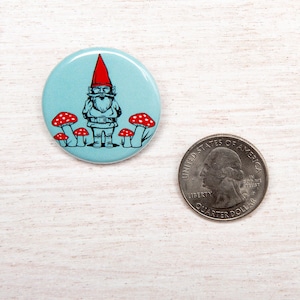 May include: A circular badge with a light blue backdrop depicts a cartoon gnome wearing a red hat and white beard, surrounded by red and white mushrooms. A US quarter is next to the badge for size comparison.
