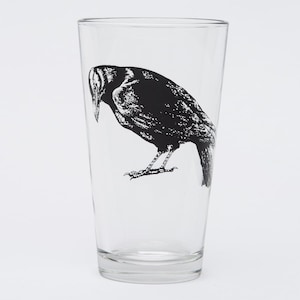 May include: A clear glass pint glass featuring a black illustration of a crow. The crow is depicted in a detailed, artistic style, with its head bowed and its body angled downward. The glass is empty and sits against a white background.