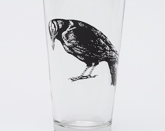 May include: A clear glass pint glass featuring a black illustration of a crow. The crow is depicted in a detailed, artistic style, with its head bowed and its body angled downward. The glass is empty and sits against a white background.
