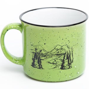 May include: A light green ceramic mug with a speckled finish and a black rim. The exterior features a black line art illustration of a mountain landscape with pine trees and a winding river. The interior of the mug is solid white.