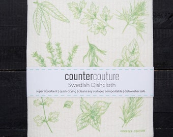 May include: A white Swedish dishcloth featuring green botanical illustrations of herbs. The dishcloth has a band with the text "countercouture Swedish Dishcloth" and phrases like "super absorbent" and "dishwasher safe".