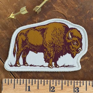 May include: Embroidered patch featuring a bison in shades of brown and burgundy, set against a light gray background. The patch is outlined with a white border. Two gold-colored arrow pins are also visible.