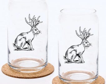 May include: Two clear glass drinking glasses with cork coasters. Each glass features a black and white illustration of a rabbit with antlers.