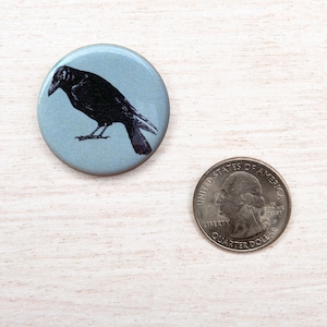 May include: A round button with a light blue background features a black illustration of a crow. The button is next to a U.S. quarter coin for size comparison. The crow is depicted in profile, with its beak and feathers detailed.