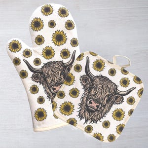 May include: An oven mitt and pot holder set. The set features a cream-colored background with a pattern of yellow sunflowers. Each item has a detailed illustration of a Highland cow's face. The oven mitt has a thumb and finger pocket.