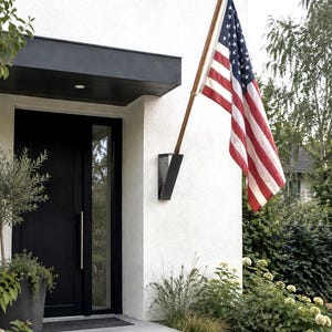 May include: A United States flag with red, white, and blue stripes and a blue field with white stars, mounted on a wooden pole attached to a black metal bracket on a white exterior wall next to a black front door with a vertical silver handle.