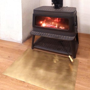May include: A black wood-burning stove with a glass door and a fan on top. The stove is sitting on a stand and has a brass fireproof mat underneath. The stove is lit and there are flames visible inside.