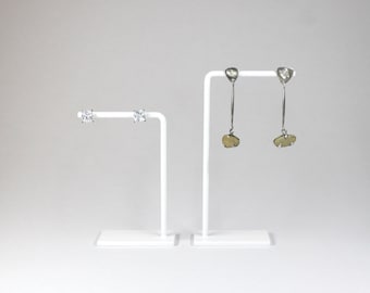 May include: Two white earring stands with earrings. The stand on the left has two small, clear, square-shaped earrings. The stand on the right has two long, silver earrings with a light brown, oval-shaped pendant.