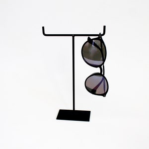 Puede incluir: Un soporte de metal negro con una base rectangular y una parte superior en forma de T. Un par de gafas de sol redondas con monturas negras y lentes oscuras cuelgan del soporte.