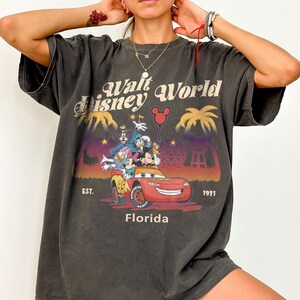 May include: Dark grey t-shirt with a colourful graphic of Disney characters in a red car, the text reads "Walt Disney World Florida" and "EST. 1971". The shirt has a vintage, worn appearance.