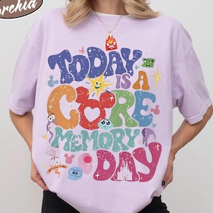 May include: A light purple Comfort Colors t-shirt with the word "orchid" and the phrase "Today is a Core Memory Day" in colorful, cartoonish lettering. The shirt features various cartoon character graphics.