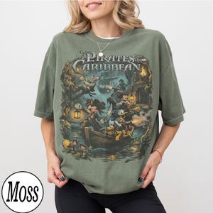May include: Khaki green t-shirt with a detailed graphic from "Pirates of the Caribbean" featuring Mickey Mouse, Donald Duck, and other characters in a pirate ship scene. The shirt has the text "Pirates of the Caribbean".