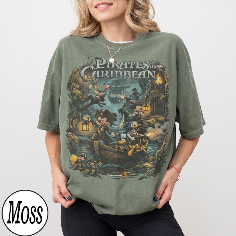 May include: Khaki green t-shirt with a detailed graphic from "Pirates of the Caribbean" featuring Mickey Mouse, Donald Duck, and other characters in a pirate ship scene. The shirt has the text "Pirates of the Caribbean".