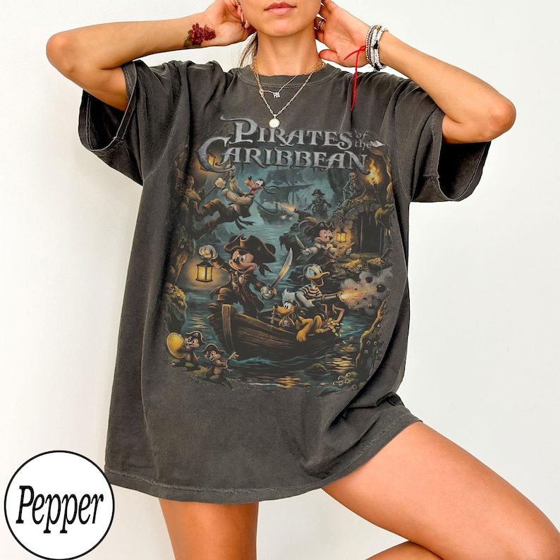 May include: Dark grey t-shirt with a Pirates of the Caribbean graphic, featuring Mickey Mouse, Donald Duck, and other Disney characters in a pirate ship. The shirt has a distressed, vintage appearance.