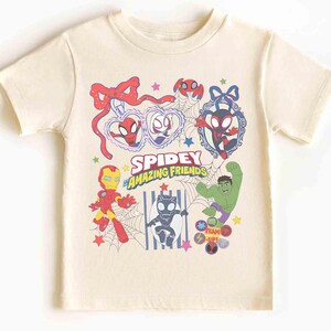 Może przedstawiać: Kremowy t-shirt z kolorową grafiką Spidey i jego Niesamowitych Przyjaci&oacute;ł. Projekt zawiera r&oacute;żne postacie, takie jak Iron Man i Hulk, wraz z tekstem "Spidey & Amazing Friends".