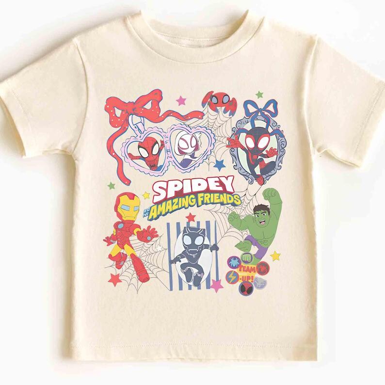 Może przedstawiać: Kremowy t-shirt z kolorową grafiką Spidey i jego Niesamowitych Przyjaci&oacute;ł. Projekt zawiera r&oacute;żne postacie, takie jak Iron Man i Hulk, wraz z tekstem "Spidey & Amazing Friends".