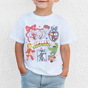 Może przedstawiać: Biała koszulka z napisem "Spidey Amazing Friends" i kresk&oacute;wkowymi ilustracjami Spider-Mana, Iron Mana, Hulka i innych postaci. Projekt zawiera pajęczyny, gwiazdy i ramkę w kształcie serca.