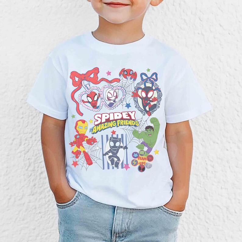 Może przedstawiać: Biała koszulka z napisem "Spidey Amazing Friends" i kresk&oacute;wkowymi ilustracjami Spider-Mana, Iron Mana, Hulka i innych postaci. Projekt zawiera pajęczyny, gwiazdy i ramkę w kształcie serca.