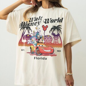 May include: Cream-coloured t-shirt with a colourful graphic of Disney characters in a red car, and the text "Walt Disney World Florida Est. 1971". The design includes palm trees and a Mickey Mouse balloon.
