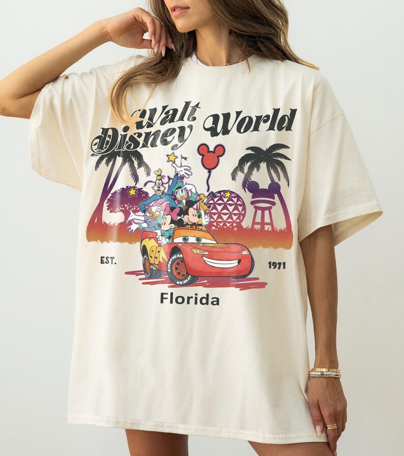 May include: Cream-coloured t-shirt with a colourful graphic of Disney characters in a red car, and the text "Walt Disney World Florida Est. 1971". The design includes palm trees and a Mickey Mouse balloon.