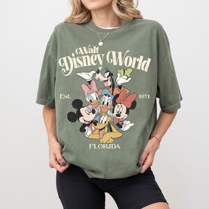 May include: Olive green t-shirt featuring a graphic of Disney characters including Mickey Mouse, Minnie Mouse, Donald Duck, and Goofy. The text "Walt Disney World" is arched above the characters, with "Est. 1971" and "Florida" below.