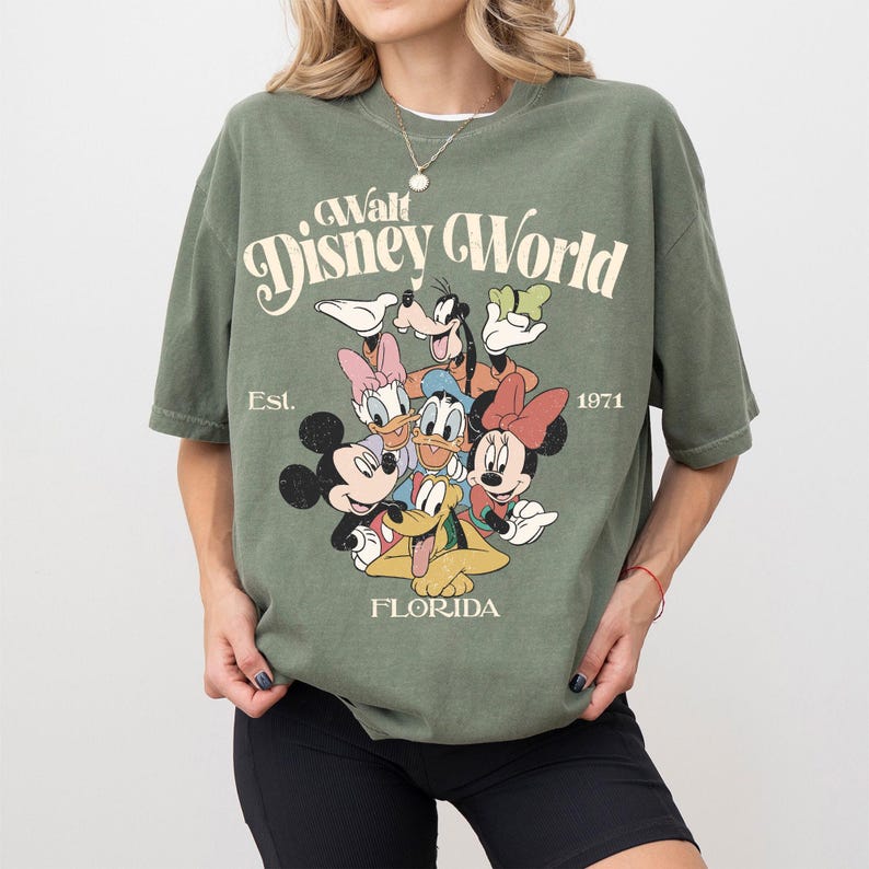 May include: Olive green t-shirt featuring a graphic of Disney characters including Mickey Mouse, Minnie Mouse, Donald Duck, and Goofy. The text "Walt Disney World" is arched above the characters, with "Est. 1971" and "Florida" below.