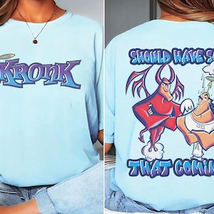 May include: Light blue t-shirt featuring the word "Kronk" in purple and blue, with a devil tail and halo. The back of the shirt displays a graphic of a red devil and an angel with the text "SHOULD HAVE SEEN THAT COMING" in blue.