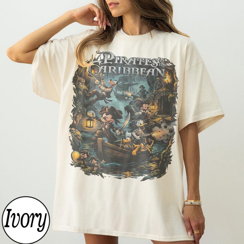May include: Ivory-coloured t-shirt with a "Pirates of the Caribbean" graphic. The design features Mickey Mouse, Donald Duck, and other characters in a pirate scene. The shirt has a loose fit.
