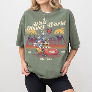 May include: An olive green t-shirt with a vintage Walt Disney World graphic. The design features cartoon characters in a red car, palm trees, and the text "Walt Disney World Florida EST. 1971".